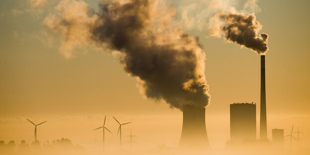 Eine Kohlekraftwerk stößt Dampf aus, dahinter Windräder, die Szenerie ist in gelbliches Licht getaucht