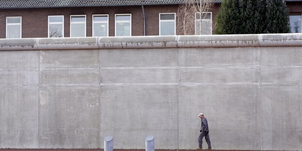 Ein Arbeiter eine hohe Mauer entlang, hinter der ein Haus und ein Baum zu sehen sind Ein Arbeiter eine hohe Mauer entlang, hinter der ein Haus und ein Baum zu sehen sind