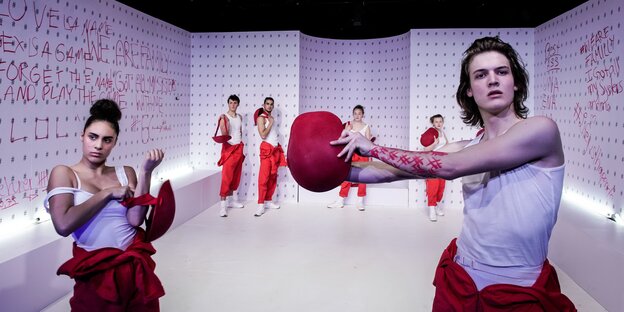 Im Vordergrund posieren zwei Schauspieler*innen, im Hintergrund stehen die anderen an der Wand. Die Wand ist vollgeschrieben mit Sätzen in roter Schrift. Im Vordergrund posieren zwei Schauspieler*innen, im Hintergrund stehen die anderen an der Wand. Die Wand ist vollgeschrieben mit Sätzen in roter Schrift.