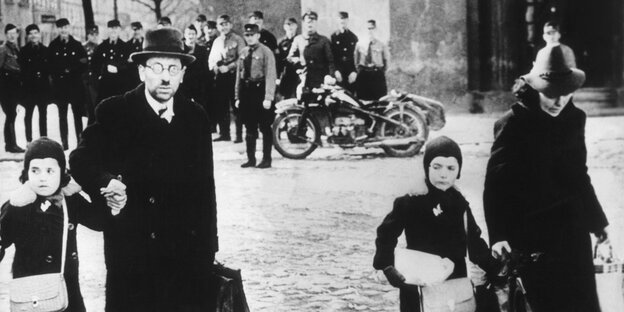 Eine Familie läuft über einen Platz, hinter ihnen stehen Männer in Uniform, manche lachen Eine Familie läuft über einen Platz, hinter ihnen stehen Männer in Uniform, manche lachen