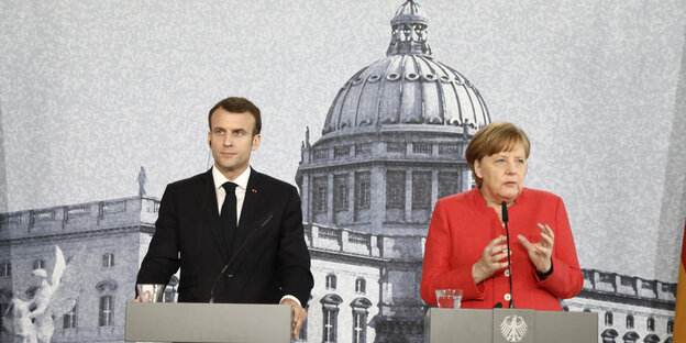 Macron im schwarzen Jackett, Merkel im roten, stehen vor einer Fototapete Macron im schwarzen Jackett, Merkel im roten, stehen vor einer Fototapete