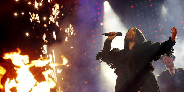 Conchita Wurst hält ein Mikrofon in der Hand und singt, neben ihr lodert ein Feuer Conchita Wurst hält ein Mikrofon in der Hand und singt, neben ihr lodert ein Feuer