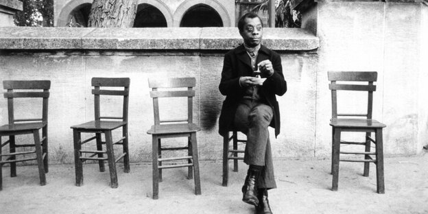 James Baldwin sitzt auf einem Stuhl, neben sich mehrere leere Stühle James Baldwin sitzt auf einem Stuhl, neben sich mehrere leere Stühle