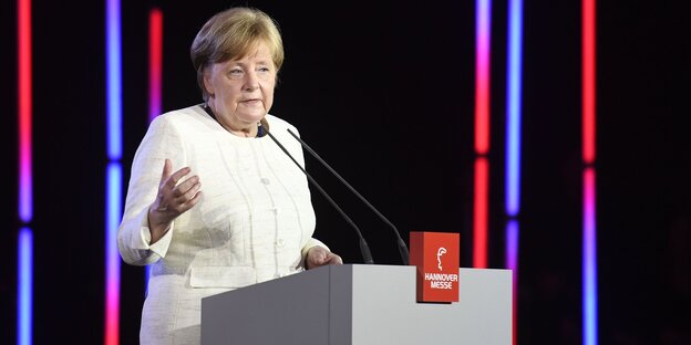 Eine Frau in weißer Bluse spricht in Mikrofone auf einem Podium, im Hintergrund bunte Lichtstreifen Eine Frau in weißer Bluse spricht in Mikrofone auf einem Podium, im Hintergrund bunte Lichtstreifen