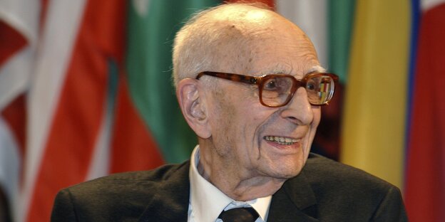 Ein alter Mann mit Glatze und Brille, im Hintergrund verschiedene Flaggen Ein alter Mann mit Glatze und Brille, im Hintergrund verschiedene Flaggen