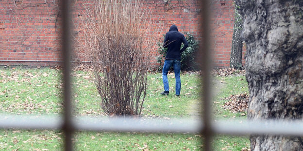 Mann in schwarzer Kaputze läuft hinter Gittern durch einen Garten Mann in schwarzer Kaputze läuft hinter Gittern durch einen Garten