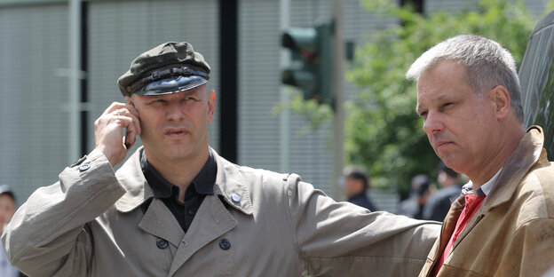 Zwei Männer stehen vor einer Kreuzung. Einer trägt Trenchcoat und Schiebermütze, der andere eine braune Jacke. Zwei Männer stehen vor einer Kreuzung. Einer trägt Trenchcoat und Schiebermütze, der andere eine braune Jacke.