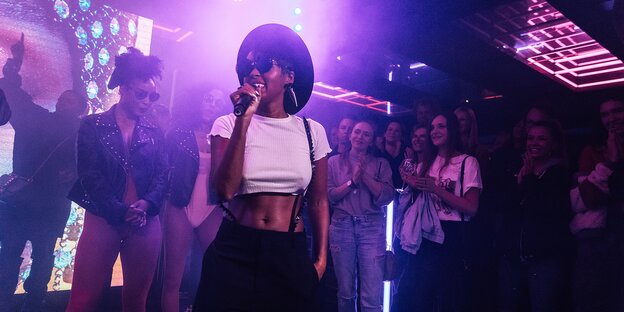 Die Künstlerin Janelle Monáe steht in lilafarbenem Licht und hält ein Mikrofon in der Hand Die Künstlerin Janelle Monáe steht in lilafarbenem Licht und hält ein Mikrofon in der Hand