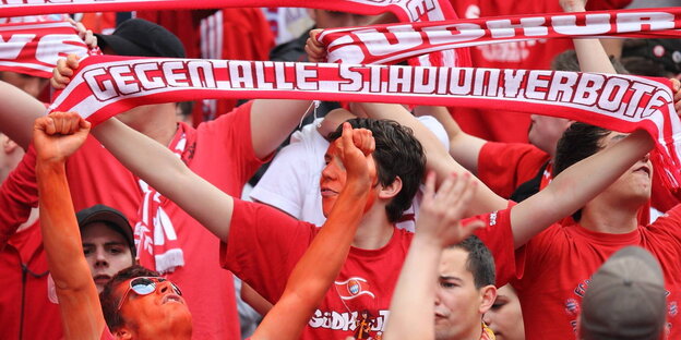 Menschen in roter Kleidung. Einer hält einen Schal mit der Aufschrift „Gegen alle Stadionverbote" Menschen in roter Kleidung. Einer hält einen Schal mit der Aufschrift „Gegen alle Stadionverbote"