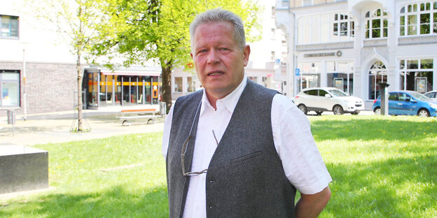 Ein älterer Mann in weißem Tshirt und grauer Weste steht auf einer grünen Wiese. Die Sonne scheint Ein älterer Mann in weißem Tshirt und grauer Weste steht auf einer grünen Wiese. Die Sonne scheint