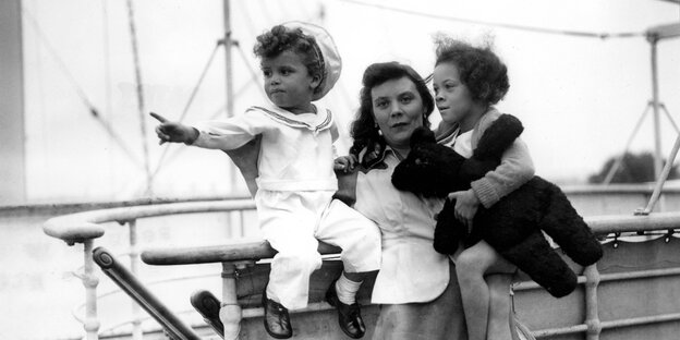 Ein Foto in schwarz weiß zeigt eine junge Frau mit einem Jungen und einem Mädchen auf dem Arm an Bord eines Schiffes. Der Junge trägt einen Matrosenanzug, das Mädchen ein Kleid und ein Plüschtier Ein Foto in schwarz weiß zeigt eine junge Frau mit einem Jungen und einem Mädchen auf dem Arm an Bord eines Schiffes. Der Junge trägt einen Matrosenanzug, das Mädchen ein Kleid und ein Plüschtier