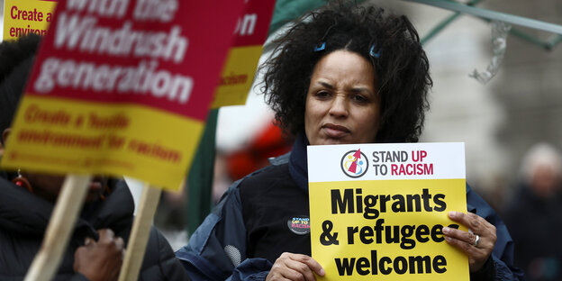 Eine schwarze Frau hält ein gelbes Schild auf dem steht: Migrants and refugees welcome here Eine schwarze Frau hält ein gelbes Schild auf dem steht: Migrants and refugees welcome here