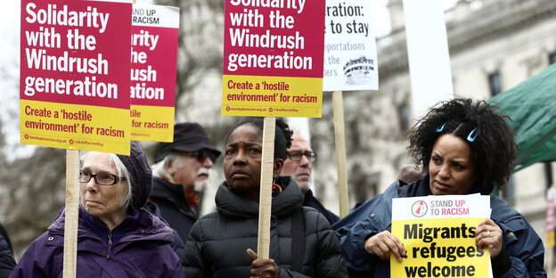 Frauen halten Schilder in die Höhe, auf denen „Solidarity with the Windrush Generation“ steht Frauen halten Schilder in die Höhe, auf denen „Solidarity with the Windrush Generation“ steht