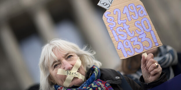 Eine Frau hat sich den Mund mit Tape verklebt, auf dem „219a“ steht. Sie hält ein Schild in der Hand mit der Aufschrift „§219a ist so 1933“ Eine Frau hat sich den Mund mit Tape verklebt, auf dem „219a“ steht. Sie hält ein Schild in der Hand mit der Aufschrift „§219a ist so 1933“