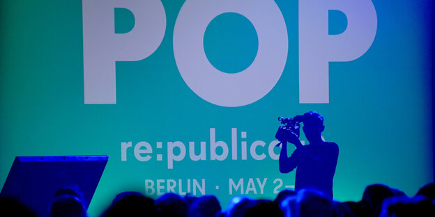Die Silouette eines Menschen, der eine Kamera hält, vor einer türkis-blauen Wand, auf der „POP“ und „re:publica“ steht Die Silouette eines Menschen, der eine Kamera hält, vor einer türkis-blauen Wand, auf der „POP“ und „re:publica“ steht
