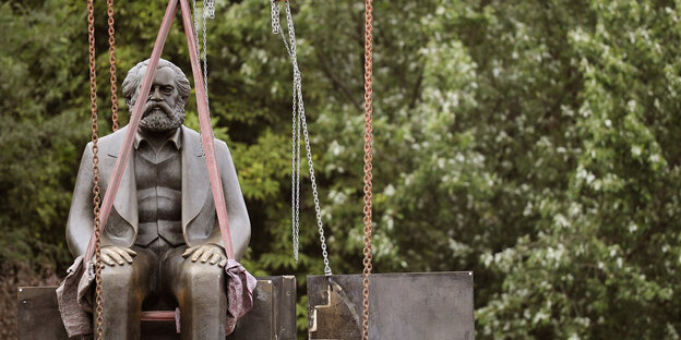 Eine Statue von einem sitzenden, älteren Mann hängt an Seilen in der Luft, im Hintergrund sieht man das Grün eines Baums. Eine Statue von einem sitzenden, älteren Mann hängt an Seilen in der Luft, im Hintergrund sieht man das Grün eines Baums.