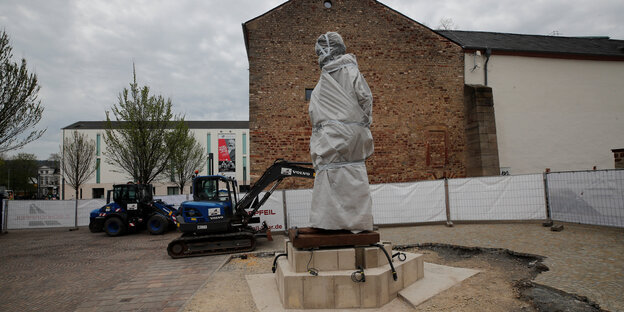 Eine in weißte Tücher eingehüllte Statue vor einer Brandwand. Der Kopf sieht durch die Verhüllung aus wie das aufgerissene Maul eines Löwen Eine in weißte Tücher eingehüllte Statue vor einer Brandwand. Der Kopf sieht durch die Verhüllung aus wie das aufgerissene Maul eines Löwen