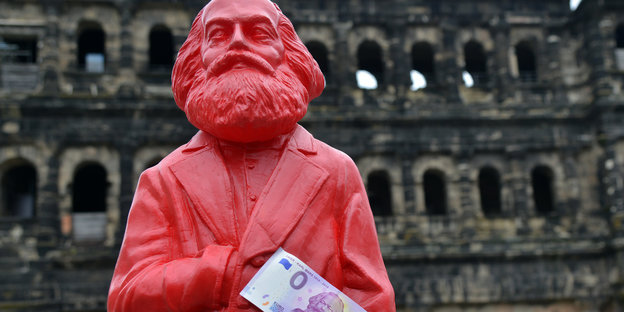 Eine rote Figur, die Karl Marx darstellt vor der Porta Nigra in Trier. An seiner hand klebt ein o-Euro-Schein