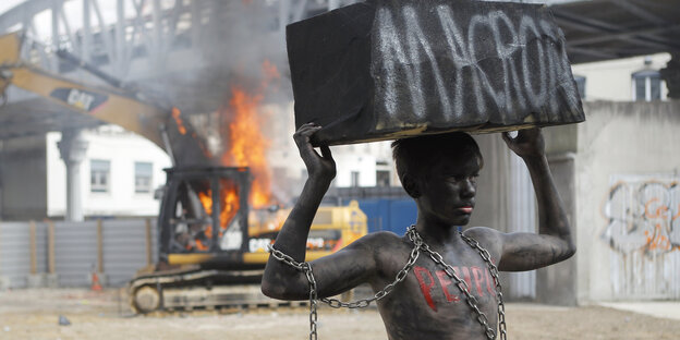 Ein junger Aktivist mit schwarz angemaltem Körper hebt einen Stein über den Kopf, auf dem "Macron" steht