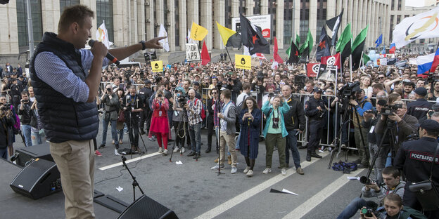Ein Mann spricht an einem Mikrofon zu einer großen Menschenmenge, die mit Bannern und Flaggen weht Ein Mann spricht an einem Mikrofon zu einer großen Menschenmenge, die mit Bannern und Flaggen weht
