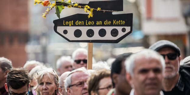 Menschen stehen nebeneinander, über ihnen ragt ein Schild in Form eines Panzers in die Höhe, auf dem „Legt den Leo an die Kette“ steht. Eine Blume ist an das Panzerrohr gebunden Menschen stehen nebeneinander, über ihnen ragt ein Schild in Form eines Panzers in die Höhe, auf dem „Legt den Leo an die Kette“ steht. Eine Blume ist an das Panzerrohr gebunden
