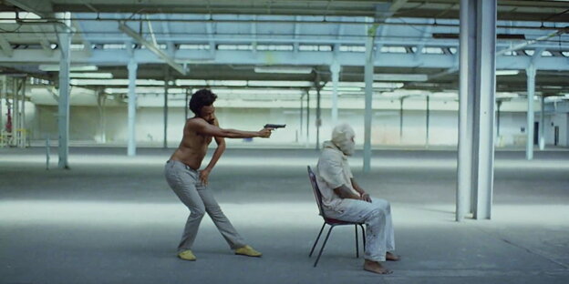 Szene aus dem Musikvideo, in dem Childish Gambino seine Waffen auf einen Mann richtet. Dieser sitzt mit Tuch über dem Kopf auf einem Stuhl. Szene aus dem Musikvideo, in dem Childish Gambino seine Waffen auf einen Mann richtet. Dieser sitzt mit Tuch über dem Kopf auf einem Stuhl.