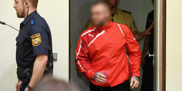 Ein Mann im roten Trainingsdress tritt hinter einem Vollzugsbeamten in den Saal, sein Gesicht ist verpixelt Ein Mann im roten Trainingsdress tritt hinter einem Vollzugsbeamten in den Saal, sein Gesicht ist verpixelt