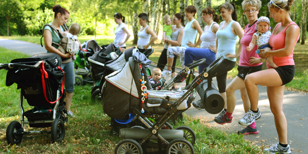 Mütter in Sportbekleidung, mit Babys auf dem Arm und mit Kinderwagen beim Sport im Park Mütter in Sportbekleidung, mit Babys auf dem Arm und mit Kinderwagen beim Sport im Park