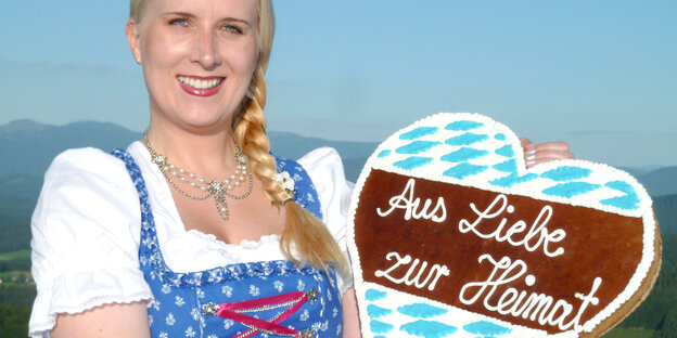 Eine Frau im Dirndl hält ein Lebkuchenherz mit der Aufschrift "Aus Liebe zur Heimat".