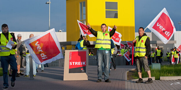 Männer in gelben Westen stehen vor einem gelben Container und haben Fahnen mit der Aufschrift verdi in der Hand Männer in gelben Westen stehen vor einem gelben Container und haben Fahnen mit der Aufschrift verdi in der Hand
