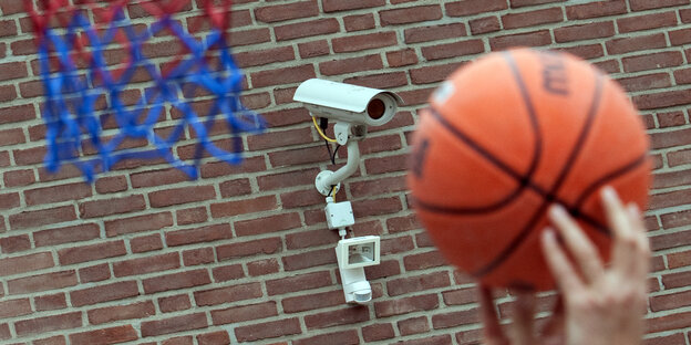 Zwei Hände, die einen Basketball halten. Im Hintergrund eine Mauer mit Überwachungskamera. Zwei Hände, die einen Basketball halten. Im Hintergrund eine Mauer mit Überwachungskamera.