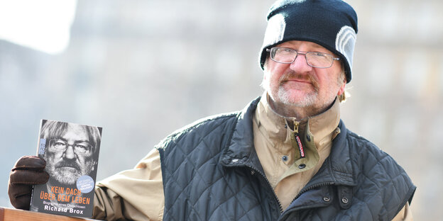 Richard Brox hat einen Arm auf die Lehne einer Bank gelegt und hält sein Buch in der Hand Richard Brox hat einen Arm auf die Lehne einer Bank gelegt und hält sein Buch in der Hand