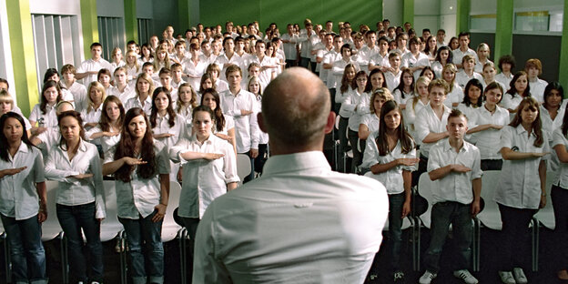 Man sieht einen Mann von hinten, der vor einer Menge junger Menschen in einheitlicher Uniform steht. Alle Schüler stehen in Reih' und Glied und halten sich die rechte Hand vor die Brust