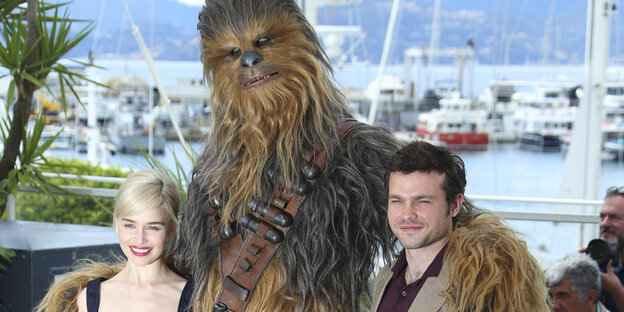 Der Schauspieler Alden Ehrenreich und die Schauspielerin Emilia Clarke stehen neben einer Person in einem Kostüm von Chewbacca Der Schauspieler Alden Ehrenreich und die Schauspielerin Emilia Clarke stehen neben einer Person in einem Kostüm von Chewbacca