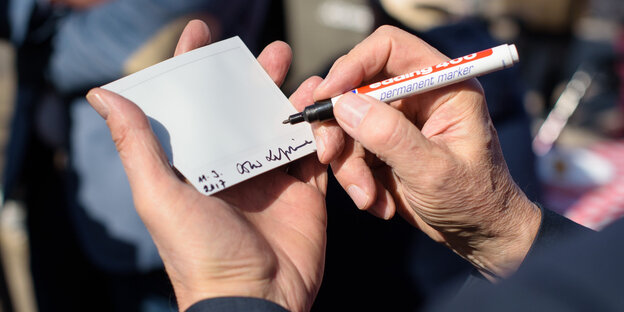 Eine Hand hält ein weißes Stück Papier, eine andere schreibt mit Edding darauf Eine Hand hält ein weißes Stück Papier, eine andere schreibt mit Edding darauf