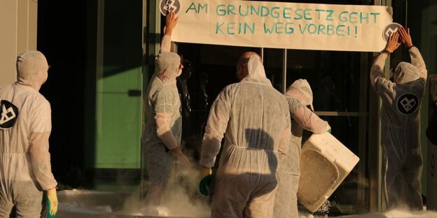 Menschen in weißen Schutzanzügen vor einer Eingangstür. Zwei von ihnen befestigen ein Transparent mit der Aufschrift „Am Grundgesetz geht kein Weg vorbei“. Auf dem Boden dampfen Eisblöcke Menschen in weißen Schutzanzügen vor einer Eingangstür. Zwei von ihnen befestigen ein Transparent mit der Aufschrift „Am Grundgesetz geht kein Weg vorbei“. Auf dem Boden dampfen Eisblöcke