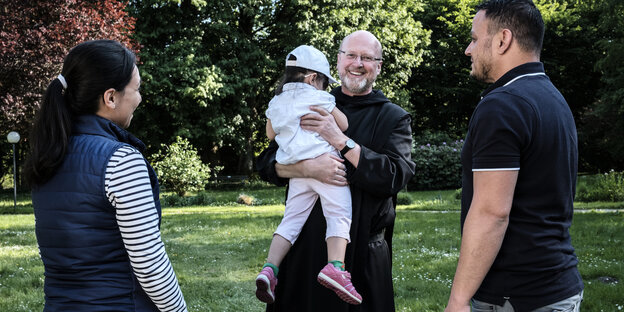 Ein Pfarrer hält ein Kind, zwei Erwachsene stehen daneben Ein Pfarrer hält ein Kind, zwei Erwachsene stehen daneben