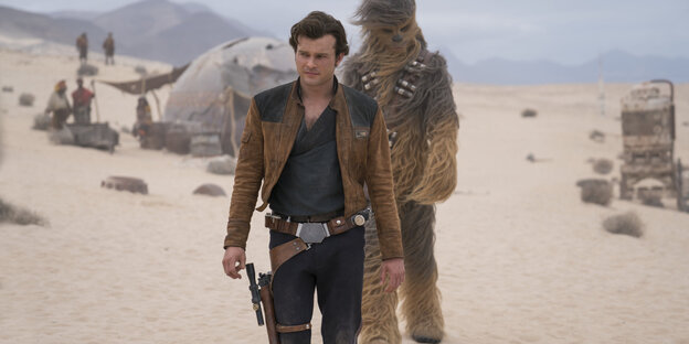 Ein Mann, es ist Han Solo, und ein Zweibeiner mit viel Fell laufen über einen windigen Wüstenplaneten Ein Mann, es ist Han Solo, und ein Zweibeiner mit viel Fell laufen über einen windigen Wüstenplaneten