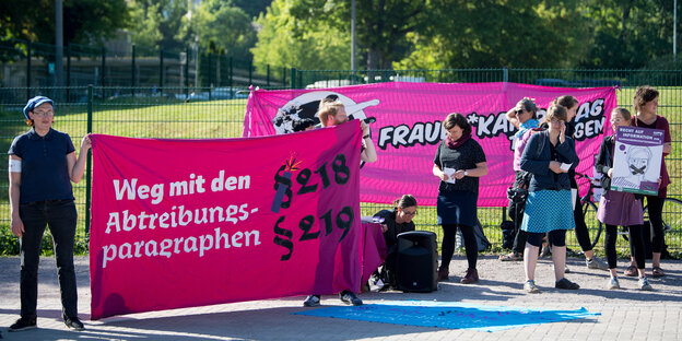 Demonstrantinnen stehen mit Plakaten vor einem Zaun Demonstrantinnen stehen mit Plakaten vor einem Zaun