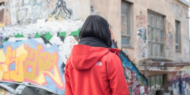 Eine Frau steht in einer roten Jacke mit dem Rücken zur Kamera vor einem Haus Eine Frau steht in einer roten Jacke mit dem Rücken zur Kamera vor einem Haus