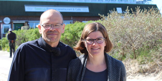 Ein Mann und eine Frau auf einer Strandpromenade - im Hintergrund ein Restaurant und eine Dünenböschung Ein Mann und eine Frau auf einer Strandpromenade - im Hintergrund ein Restaurant und eine Dünenböschung
