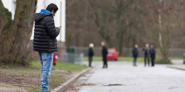 Ein Mann steht mit Smartphone am Wegesrand, weiter hinten sind unscharf weitere Personen zu sehen. Ein Mann steht mit Smartphone am Wegesrand, weiter hinten sind unscharf weitere Personen zu sehen.