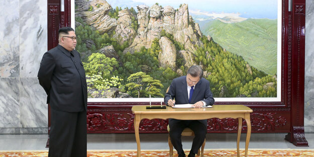 Vor einem großen Bild steht Kim Jong Un, an dem Tisch daneben sitzt Südkoreas Präsident und schreibt sich in das Gästebuch ein. Vor einem großen Bild steht Kim Jong Un, an dem Tisch daneben sitzt Südkoreas Präsident und schreibt sich in das Gästebuch ein.