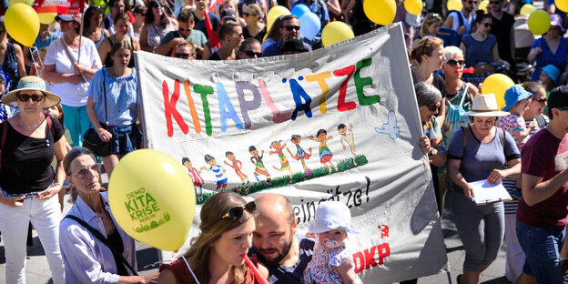 Viele Menschen um ein Transparent herum, auf dem mehr Kitaplätze gefordert werden Viele Menschen um ein Transparent herum, auf dem mehr Kitaplätze gefordert werden