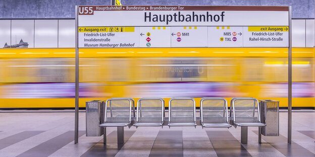 U-Bahnstation, im Mittelpunkt eine Sitzreihe, dahinter ein gelber U-Bahnzug, der schnell ein- oder abfährt