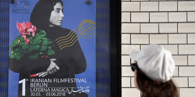 Man sieht das Plakat des 1. Iranischen Filmfestivals, darauf ist eine Frau in schwarz zu sehen, die einen Blumenstrauß trägt, der Hintergrund ist blau. Darunter stehen die Daten (30. Mai bis 3. Jun). EIne Frau schaut auf das Plakat Man sieht das Plakat des 1. Iranischen Filmfestivals, darauf ist eine Frau in schwarz zu sehen, die einen Blumenstrauß trägt, der Hintergrund ist blau. Darunter stehen die Daten (30. Mai bis 3. Jun). EIne Frau schaut auf das Plakat