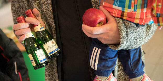 Eine Person hält zwei Flaschen Sekt und einen Apfel in der Hand. Im Arm hat sie ein Kind Eine Person hält zwei Flaschen Sekt und einen Apfel in der Hand. Im Arm hat sie ein Kind