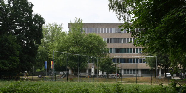 Ein braun-grauer Flachbau hinter Bäumen Ein braun-grauer Flachbau hinter Bäumen