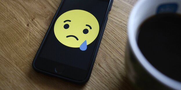 Ein Facebook-Smiley mit Tränen auf einem Smarphone neben einer Kaffeetasse Ein Facebook-Smiley mit Tränen auf einem Smarphone neben einer Kaffeetasse