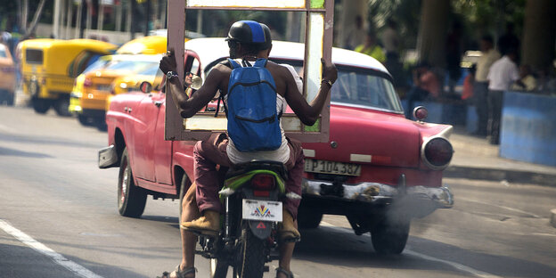 Zwei Männer auf einem Motorrad transporieren einen Bilderrahmen in Kubas Hauptstadt Havanna. Zwei Männer auf einem Motorrad transporieren einen Bilderrahmen in Kubas Hauptstadt Havanna.